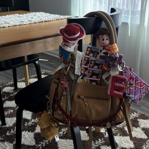May include: A tan leather handbag adorned with various accessories including two plush cowboy figures, a photo strip, a red letter E charm, a purple patterned pouch, and a red beaded garland with small metal charms hanging from the front zipper.