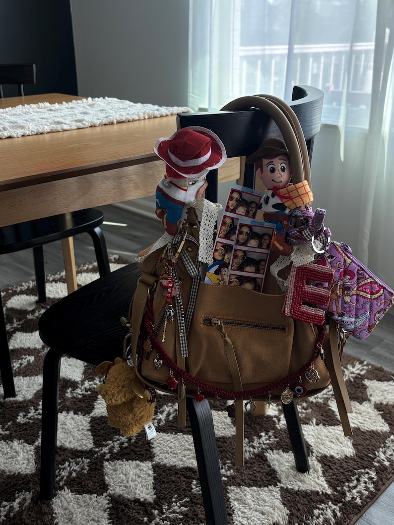 May include: A tan leather handbag adorned with various accessories including two plush cowboy figures, a photo strip, a red letter E charm, a purple patterned pouch, and a red beaded garland with small metal charms hanging from the front zipper.