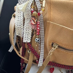 May include: A tan suede handbag featuring a gold-toned metal carabiner clip adorned with a decorative charm cluster. The charms include a small cowboy figure, red and clear beads, a cow-print bead, and letter beads spelling HOWDY, accented with brown gingham ribbon and white lace.