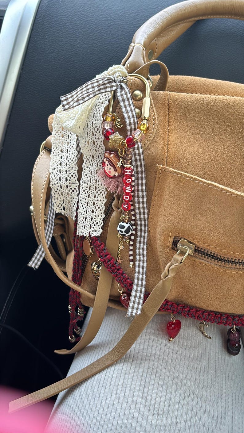 May include: A tan suede handbag featuring a gold-toned metal carabiner clip adorned with a decorative charm cluster. The charms include a small cowboy figure, red and clear beads, a cow-print bead, and letter beads spelling HOWDY, accented with brown gingham ribbon and white lace.