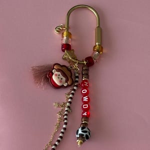 May include: A gold-toned metal carabiner keychain featuring a variety of charms including a small character wearing a brown hat, a pink tassel, and red beads spelling out the word HOWDY. Additional accents include star charms, brown and white striped beads, and a black and white cow-print bead.