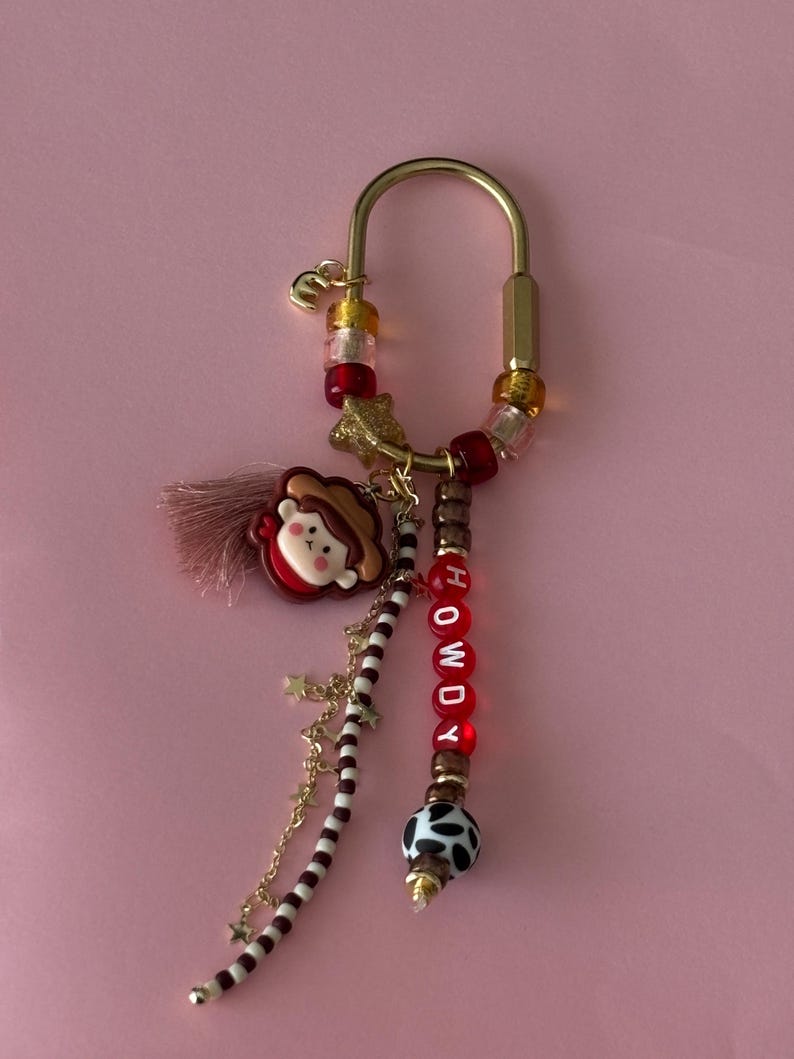May include: A gold-toned metal carabiner keychain featuring a variety of charms including a small character wearing a brown hat, a pink tassel, and red beads spelling out the word HOWDY. Additional accents include star charms, brown and white striped beads, and a black and white cow-print bead.