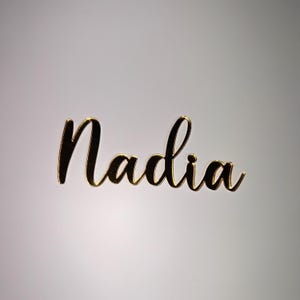 May include: Gold-colored cursive nameplate that reads "Nadia". The name is in a stylish script font with a reflective, metallic finish. The nameplate is likely used for personalized decor or gifts.