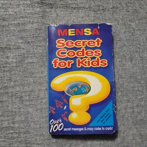 May include: A book titled "MENSA Secret Codes for Kids" with a blue cover and yellow question mark graphic. The book promises over 100 secret messages and codes to crack. The book is on a gray textured surface.