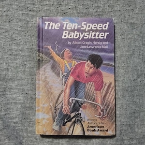 May include: A book cover with a colorful illustration of two children riding a bicycle on a beach. The book title is "The Ten-Speed Babysitter" by Alison Cragin Herzig and Jane Lawrence Mali. The book cover also includes the text "Winning Authors of the American Book Award."