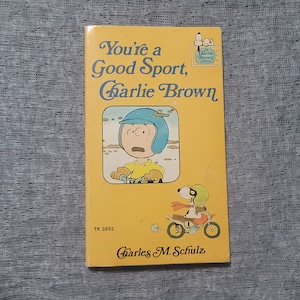 May include: A vintage paperback book titled "You're a Good Sport, Charlie Brown" with a yellow cover. The cover features an illustration of Charlie Brown on a motorcycle and Snoopy. The author's name, Charles M. Schulz, is printed at the bottom.