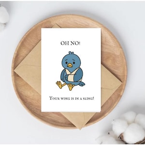 May include: A greeting card with a cartoon bird wearing a sling, with the text "OH NO! Your wing is in a sling!" The card is on a wooden plate with a brown envelope.