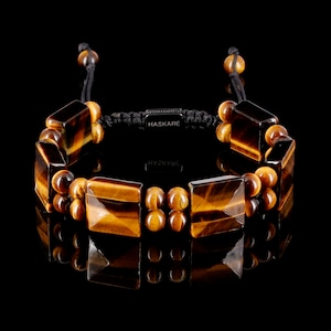 Op de afbeelding: Een bruine en gouden tijgeroog-parel armband met een zwart koord en een zilveren tag met de tekst "HASKARE".