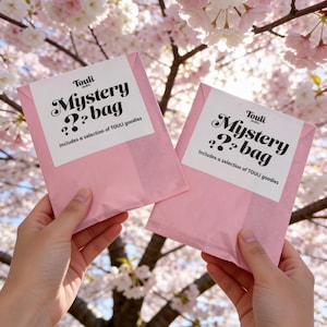 Puede incluir: Dos paquetes rosas de "Mystery bag" con etiquetas blancas, sostenidos frente a un cerezo en flor. Las etiquetas dicen "Touli Mystery bag" y "Includes a selection of TOULI goodies."