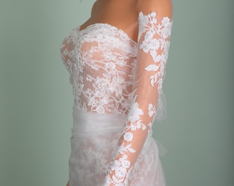 May include: A white strapless wedding dress with floral lace detailing. The dress features a fitted bodice and a flowing skirt. Sheer, long sleeves with matching floral lace complete the bridal look.