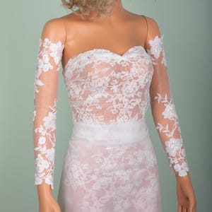Peut inclure: Robe de mari&eacute;e en dentelle blanche sans bretelles avec de longues manches transparentes. La robe pr&eacute;sente des d&eacute;tails de dentelle florale et un corsage ajust&eacute;. La robe est une robe de mari&eacute;e.