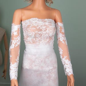 Puede incluir: Un vestido de novia blanco sin tirantes con detalles de encaje floral. El vestido presenta mangas largas y transparentes con pu&ntilde;os de encaje a juego. El vestido se exhibe en un maniqu&iacute;.