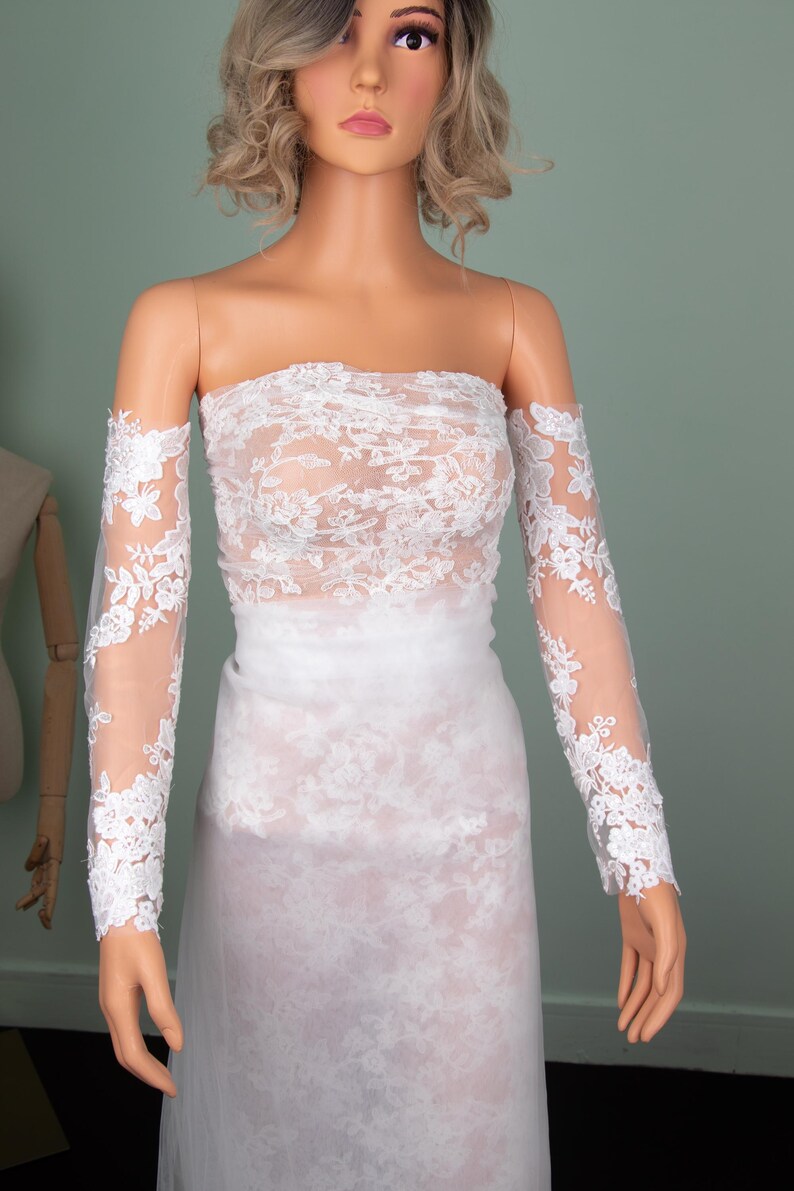 Puede incluir: Un vestido de novia blanco sin tirantes con detalles de encaje floral. El vestido presenta mangas largas y transparentes con pu&ntilde;os de encaje a juego. El vestido se exhibe en un maniqu&iacute;.