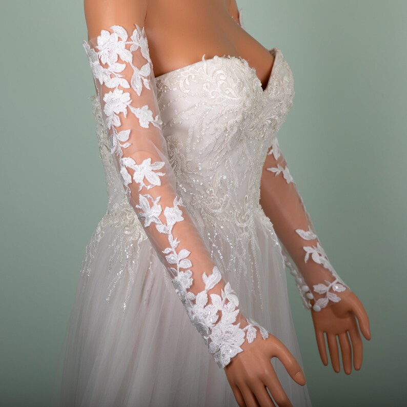 May include: A white wedding dress with a sweetheart neckline and a tulle skirt. The dress features intricate beading and lace details. Sheer, long sleeves with floral lace appliques are attached to the dress.