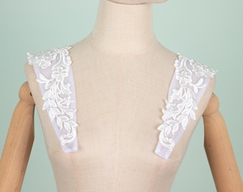 May include: White floral lace shoulder appliques. The appliques feature a delicate white lace with a floral and leaf design. They are displayed on a mannequin, highlighting their shape and detail. Ideal for bridal or formal wear.