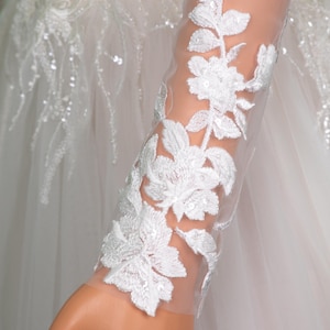 May include: Close-up of a white wedding dress sleeve with intricate floral embroidery. The delicate design features white flowers and leaves, embellished with sequins, on a sheer fabric. The dress is a classic bridal design.