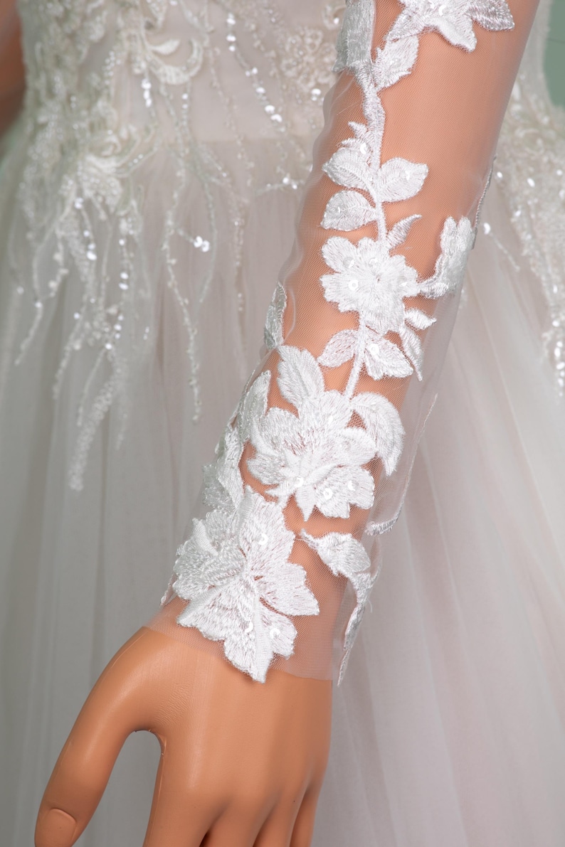 May include: Close-up of a white wedding dress sleeve with intricate floral embroidery. The delicate design features white flowers and leaves, embellished with sequins, on a sheer fabric. The dress is a classic bridal design.