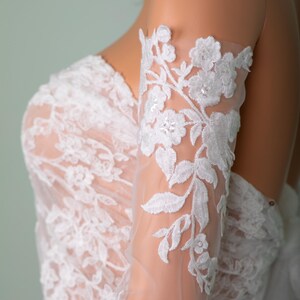 Peut inclure: Robe de mari&eacute;e en dentelle blanche avec broderie florale. Les manches transparentes pr&eacute;sentent des motifs floraux d&eacute;licats et sont orn&eacute;es de sequins. Le corsage est &eacute;galement orn&eacute; de dentelle, cr&eacute;ant un look de mari&eacute;e &eacute;l&eacute;gant.