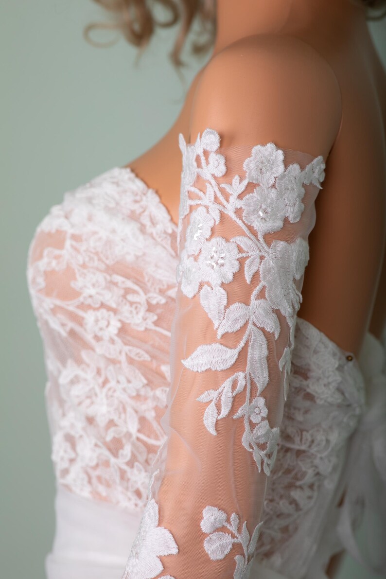 Peut inclure: Robe de mari&eacute;e en dentelle blanche avec broderie florale. Les manches transparentes pr&eacute;sentent des motifs floraux d&eacute;licats et sont orn&eacute;es de sequins. Le corsage est &eacute;galement orn&eacute; de dentelle, cr&eacute;ant un look de mari&eacute;e &eacute;l&eacute;gant.