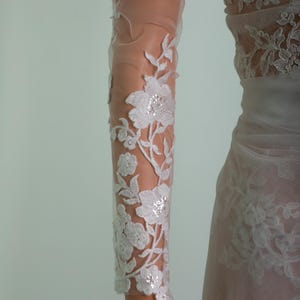 Peut inclure: Gros plan sur une manche de robe de mari&eacute;e blanche avec des d&eacute;tails de dentelle florale. Le tissu transparent r&eacute;v&egrave;le le bras en dessous, avec la dentelle pr&eacute;sentant des motifs d&eacute;licats de fleurs et de feuilles. La robe semble &ecirc;tre une robe de mari&eacute;e.