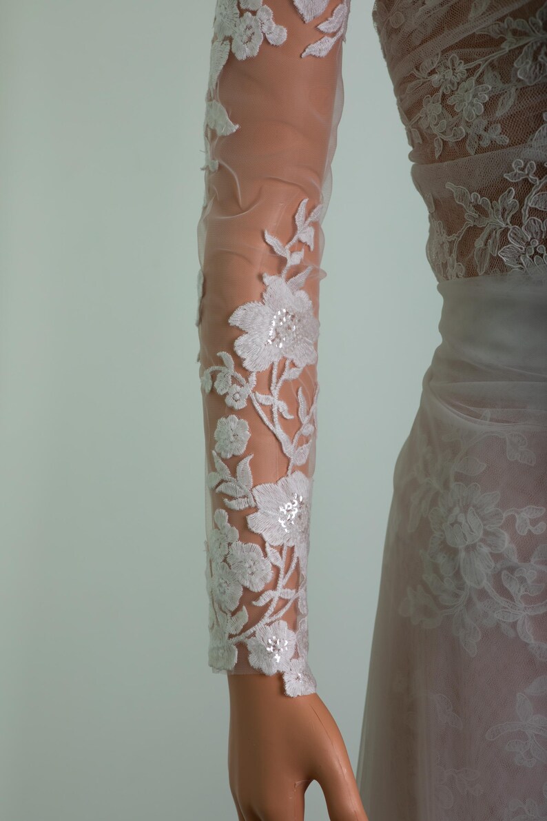 Peut inclure: Gros plan sur une manche de robe de mari&eacute;e blanche avec des d&eacute;tails de dentelle florale. Le tissu transparent r&eacute;v&egrave;le le bras en dessous, avec la dentelle pr&eacute;sentant des motifs d&eacute;licats de fleurs et de feuilles. La robe semble &ecirc;tre une robe de mari&eacute;e.