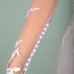 May include: Close-up of a bridal gown sleeve. The sheer fabric is adorned with white floral embroidery and small, white buttons. The sleeve is long and fitted, with a delicate, romantic design.