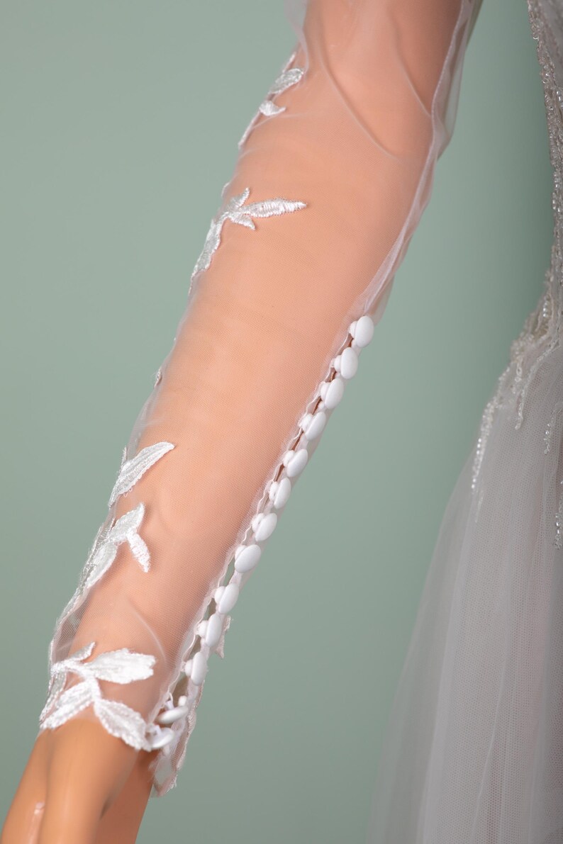 May include: Close-up of a bridal gown sleeve. The sheer fabric is adorned with white floral embroidery and small, white buttons. The sleeve is long and fitted, with a delicate, romantic design.