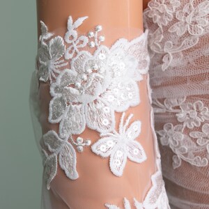 Puede incluir: Primer plano de detalles de encaje floral blanco sobre una tela transparente. El encaje presenta intrincados dise&ntilde;os de flores y hojas con detalles de lentejuelas. La tela parece ser parte de una prenda de novia o de vestir formal.