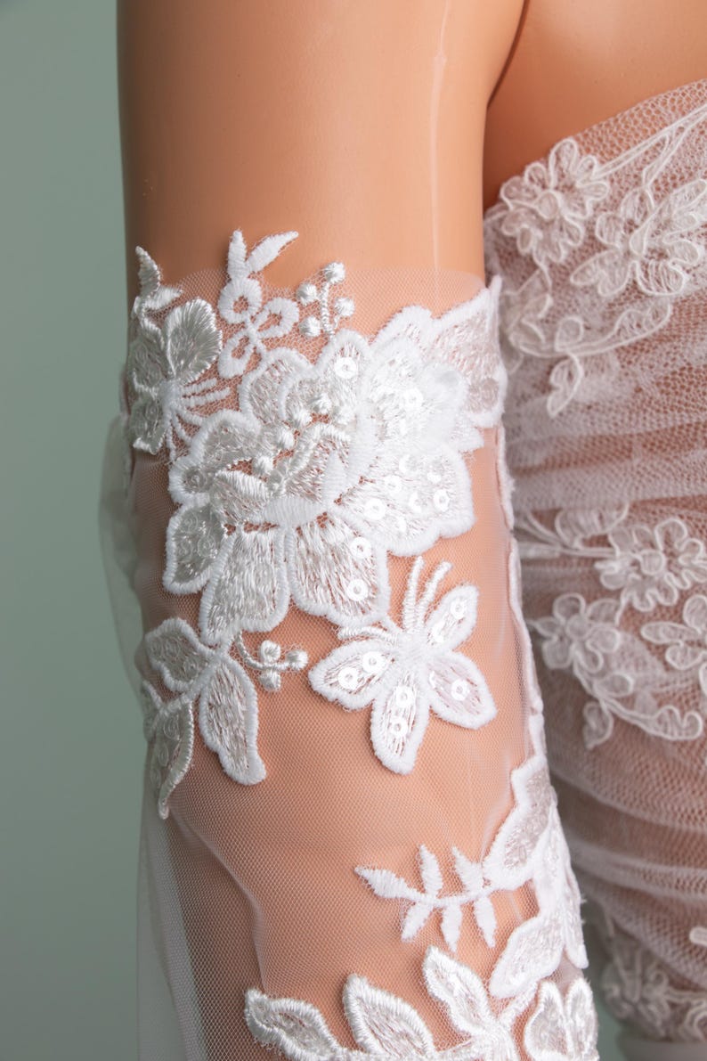 Puede incluir: Primer plano de detalles de encaje floral blanco sobre una tela transparente. El encaje presenta intrincados dise&ntilde;os de flores y hojas con detalles de lentejuelas. La tela parece ser parte de una prenda de novia o de vestir formal.