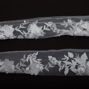 Peut inclure: Deux morceaux de garniture en dentelle florale blanche. La garniture pr&eacute;sente des fleurs et des feuilles blanches d&eacute;taill&eacute;es avec de petits embellissements scintillants. La dentelle est pos&eacute;e sur un fond noir, soulignant le design d&eacute;licat.