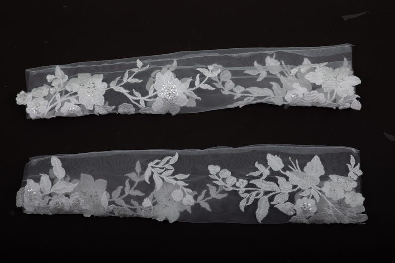 Peut inclure: Deux morceaux de garniture en dentelle florale blanche. La garniture pr&eacute;sente des fleurs et des feuilles blanches d&eacute;taill&eacute;es avec de petits embellissements scintillants. La dentelle est pos&eacute;e sur un fond noir, soulignant le design d&eacute;licat.