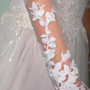 May include: Close-up of a white wedding dress sleeve with intricate floral lace detailing. The sheer fabric reveals the arm beneath, while the lace features delicate leaves and flower patterns, creating an elegant and romantic look.