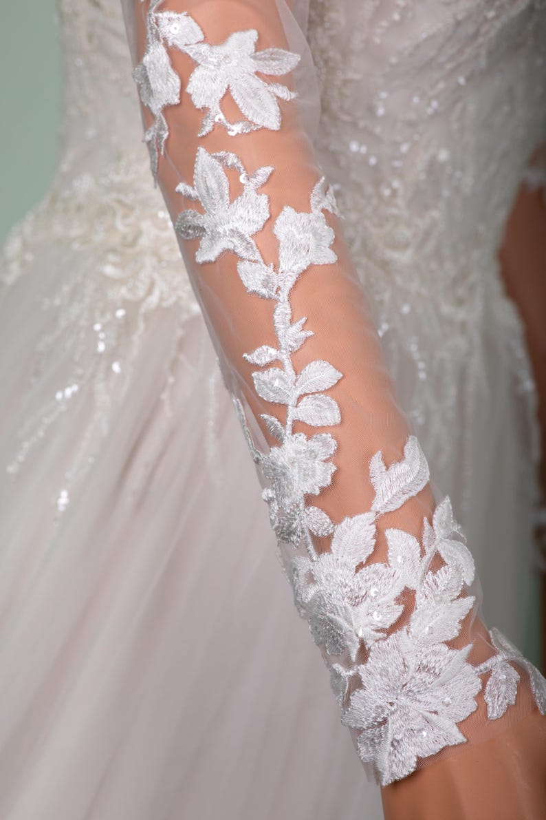May include: Close-up of a white wedding dress sleeve with intricate floral lace detailing. The sheer fabric reveals the arm beneath, while the lace features delicate leaves and flower patterns, creating an elegant and romantic look.