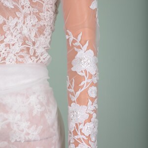 Peut inclure: Robe de mari&eacute;e en dentelle blanche avec broderie florale. La robe pr&eacute;sente une manche longue et transparente avec des motifs floraux d&eacute;taill&eacute;s et des accents de sequins. Le corsage et la jupe sont &eacute;galement orn&eacute;s de dentelle d&eacute;licate, cr&eacute;ant un look &eacute;l&eacute;gant et romantique.
