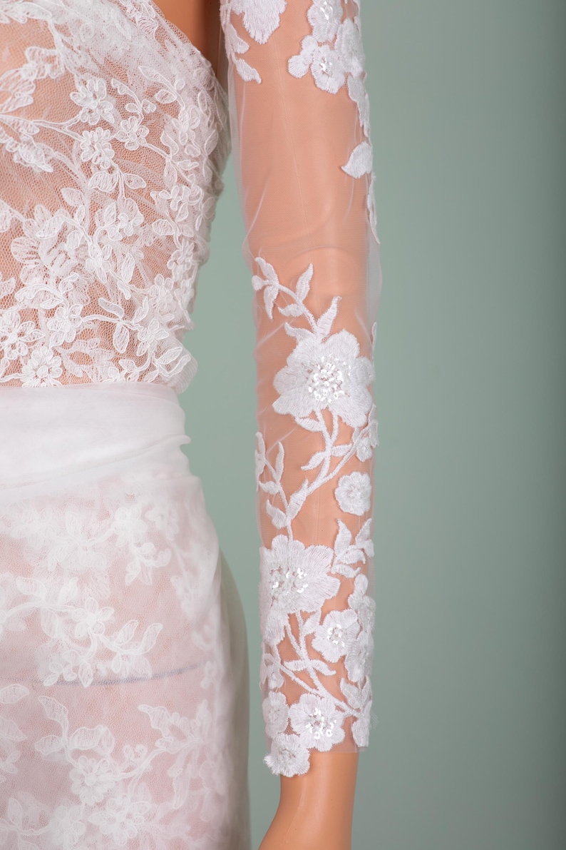 Peut inclure: Robe de mari&eacute;e en dentelle blanche avec broderie florale. La robe pr&eacute;sente une manche longue et transparente avec des motifs floraux d&eacute;taill&eacute;s et des accents de sequins. Le corsage et la jupe sont &eacute;galement orn&eacute;s de dentelle d&eacute;licate, cr&eacute;ant un look &eacute;l&eacute;gant et romantique.