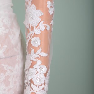 Peut inclure: Gros plan sur une manche en dentelle blanche avec broderie florale. Le motif d&eacute;licat pr&eacute;sente des fleurs et des feuilles blanches d&eacute;taill&eacute;es, avec un tissu l&eacute;ger et transparent. La manche fait partie d'une robe blanche, probablement de mari&eacute;e ou de soir&eacute;e.