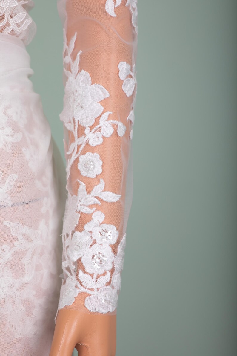 Peut inclure: Gros plan sur une manche en dentelle blanche avec broderie florale. Le motif d&eacute;licat pr&eacute;sente des fleurs et des feuilles blanches d&eacute;taill&eacute;es, avec un tissu l&eacute;ger et transparent. La manche fait partie d'une robe blanche, probablement de mari&eacute;e ou de soir&eacute;e.