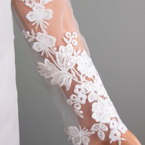 Puede incluir: Primer plano de una manga de vestido de novia blanco. La tela transparente est&aacute; adornada con intrincados bordados florales blancos. El dise&ntilde;o presenta flores y hojas detalladas, creando un aspecto delicado y elegante. La manga es larga y ajustada.