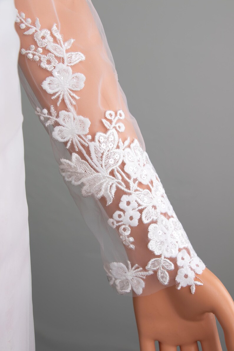 Puede incluir: Primer plano de una manga de vestido de novia blanco. La tela transparente est&aacute; adornada con intrincados bordados florales blancos. El dise&ntilde;o presenta flores y hojas detalladas, creando un aspecto delicado y elegante. La manga es larga y ajustada.