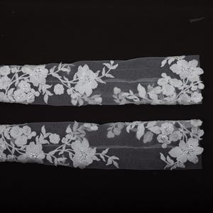 Peut inclure: Deux appliques en dentelle florale blanche avec un motif d&eacute;licat de fleurs et de feuilles. Les appliques sont orn&eacute;es de petits accents scintillants. La dentelle est pos&eacute;e sur un fond noir, soulignant les d&eacute;tails complexes.