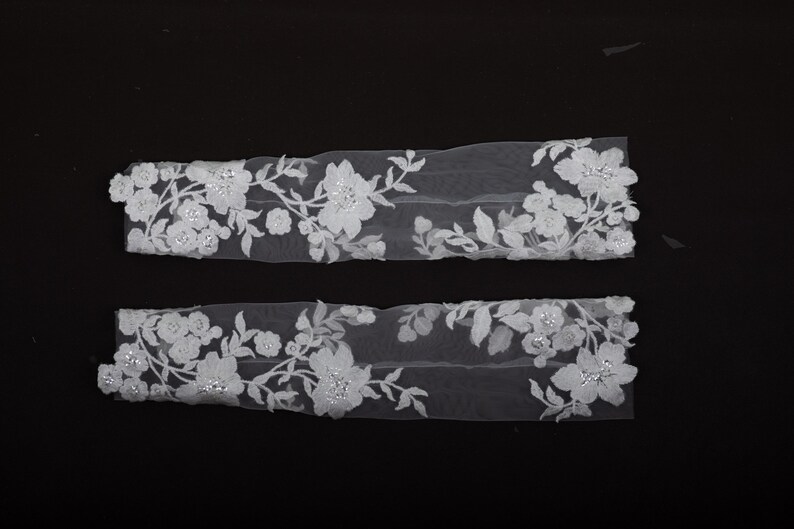 Peut inclure: Deux appliques en dentelle florale blanche avec un motif d&eacute;licat de fleurs et de feuilles. Les appliques sont orn&eacute;es de petits accents scintillants. La dentelle est pos&eacute;e sur un fond noir, soulignant les d&eacute;tails complexes.