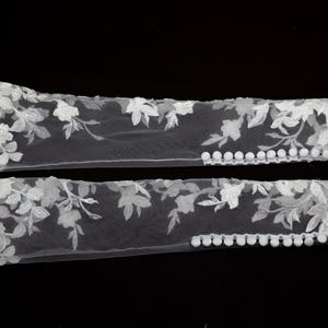 May include: Two white floral lace arm cuffs. Each cuff features delicate embroidered flowers and leaves, with small white pom-poms along one edge. The cuffs are displayed against a black background.