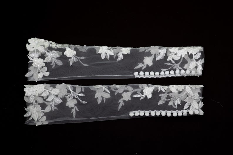 May include: Two white floral lace arm cuffs. Each cuff features delicate embroidered flowers and leaves, with small white pom-poms along one edge. The cuffs are displayed against a black background.