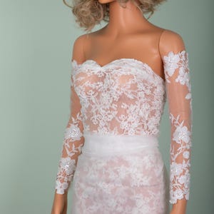 Peut inclure: Robe de mari&eacute;e en dentelle blanche avec un d&eacute;collet&eacute; &eacute;paules nues et de longues manches transparentes. La robe pr&eacute;sente des d&eacute;tails de dentelle florale complexes et un corsage ajust&eacute;. La jupe est une conception longue et fluide.