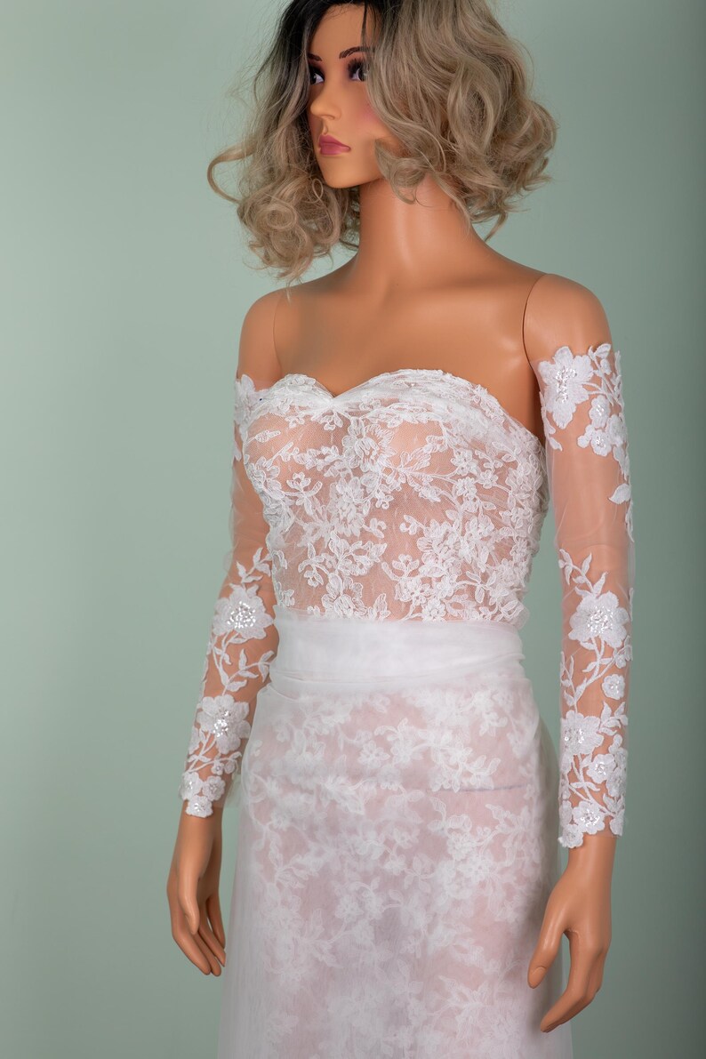 Peut inclure: Robe de mari&eacute;e en dentelle blanche avec un d&eacute;collet&eacute; &eacute;paules nues et de longues manches transparentes. La robe pr&eacute;sente des d&eacute;tails de dentelle florale complexes et un corsage ajust&eacute;. La jupe est une conception longue et fluide.