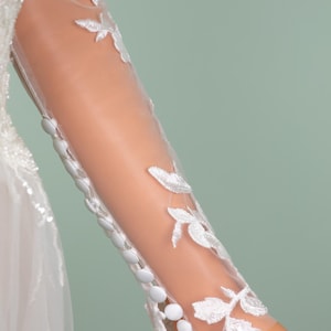 May include: Close-up of a white wedding dress sleeve. The sheer fabric is adorned with delicate white floral embroidery. A row of small, round buttons runs down the sleeve's edge. The background is a soft, muted green.