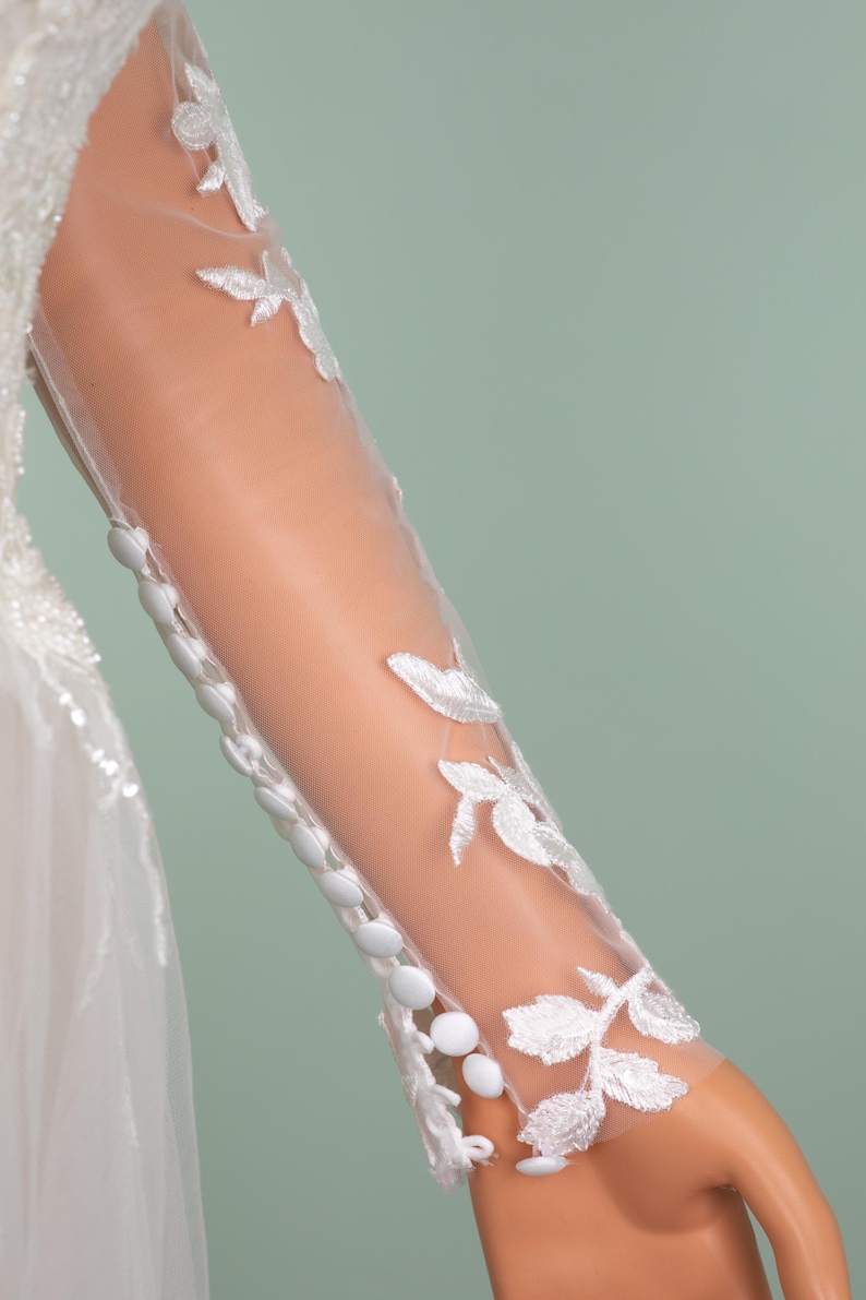 May include: Close-up of a white wedding dress sleeve. The sheer fabric is adorned with delicate white floral embroidery. A row of small, round buttons runs down the sleeve's edge. The background is a soft, muted green.