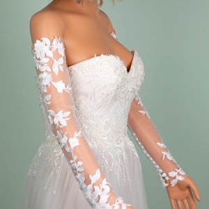 May include: A white strapless wedding dress with floral lace sleeves. The bodice is embellished with intricate beadwork and the skirt is made of a flowing tulle fabric. The sleeves are sheer, with delicate white floral lace appliqu&eacute;s.
