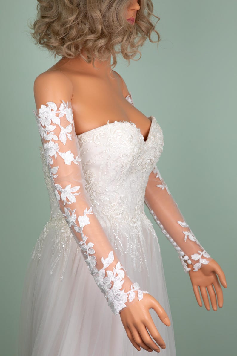 May include: A white strapless wedding dress with floral lace sleeves. The bodice is embellished with intricate beadwork and the skirt is made of a flowing tulle fabric. The sleeves are sheer, with delicate white floral lace appliqu&eacute;s.