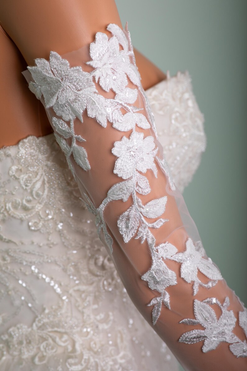 May include: Close-up of a white wedding dress sleeve with floral lace detailing. The sheer fabric is adorned with embroidered flowers and leaves, creating an elegant and delicate design. The dress features intricate beadwork.